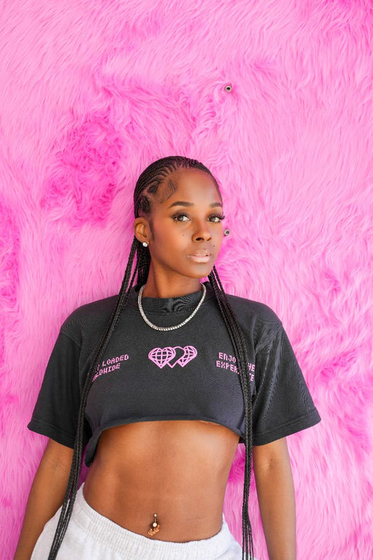 A young woman with braided hair stands against a bright pink furry backdrop in a Vintage Grey shirt