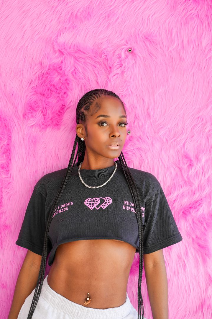 A young woman with braided hair stands against a bright pink furry backdrop in a Vintage Grey shirt